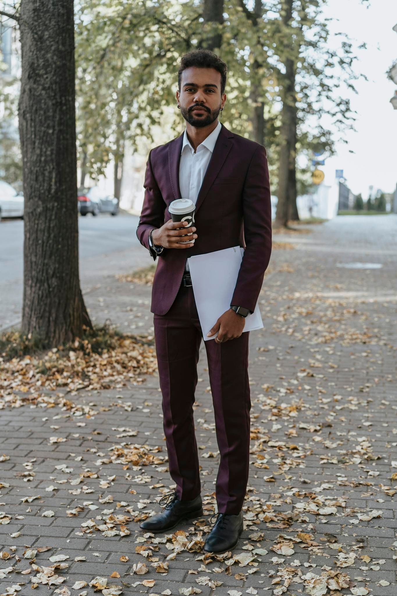 Man in suit holding coffee and document on a leaf-covered street during autumn.
